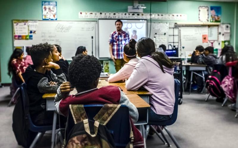 Young students sit at grouped tables in a classroom while a teacher is giving instruction in front of a whiteboard.