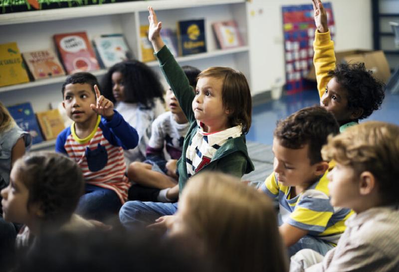 Young students sit at grouped tables in a classroom while a teacher is giving instruction in front of a whiteboard.